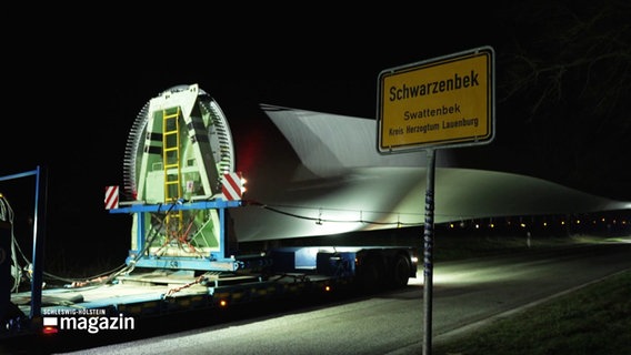 Ein riesiges Rotorblatt eines Windkraftwerks wird durch einen kleinen Ort bei Nacht transportiert. © Screenshot 