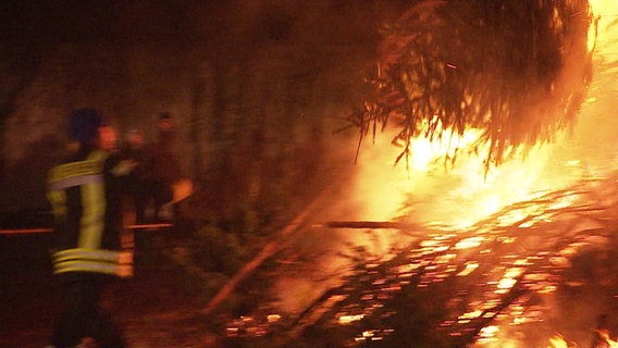 Auf einem unscharfen Bild wirft ein Feuerwehrmann einen Tannenbaum in ein größeres Feuer © Screenshot 