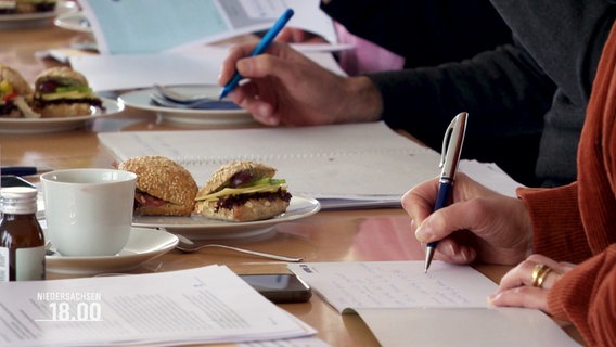 Menschen an einem Konferenztisch, auf dem Unterlagen, Kaffeetassen und Brötchen liegen- mehrere Personen schreiben. © Screenshot 