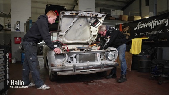 Jörg Lammers und sein Sohn stehen links und rechts von einem Oldtimer, dessen Motorhaube geöffnet ist und beugen sich über den Motorraum. © Screenshot 