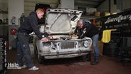 Jörg Lammers und sein Sohn stehen links und rechts von einem Oldtimer, dessen Motorhaube geöffnet ist und beugen sich über den Motorraum. © Screenshot 