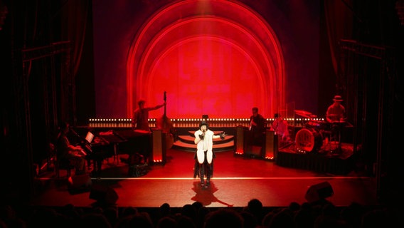 Eine Frau in einem 20er-Jahre Outfit - in schwarz, weiß mit Glitzer - steht auf einer Bühne. Rechts und links neben ihr spielt eine fünfköpfige Band © Screenshot 