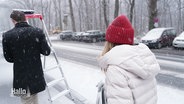 Eine Frau mit roter Mütze läuft ienem Mann mit Leiter hinterher durch den Schnee. Sie trägt ein Wahlplakat. © Screenshot 