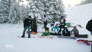 Eine Gruppe Snowboarder und Snowboarderinnen rastet auf der Pist vor einer dick verschneiten Tanne. © Screenshot 