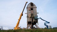 Neben einer historischen Bockwindmühle stehen ein gelber Spezialkran und eine blaue Hebebühne mit Fahrkorb. © Screenshot 