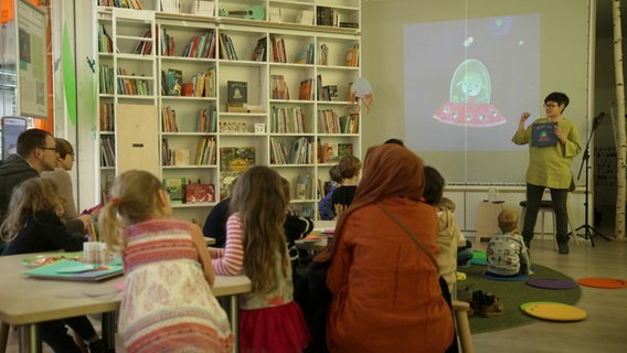 Eine Frau steht vor einer Leinwand, vor ihr sitzen viele kleine Kinder und hören ihr zu. Neben ihnen steht ein großes weißes Bücherregal voller Bücher. © Screenshot 