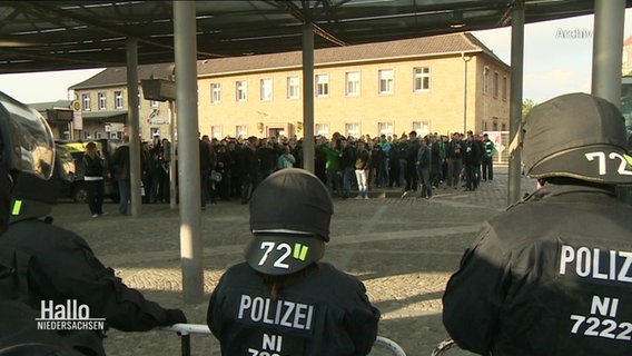 Einsatzkräfte der Polizei stehen bei einem Fan-Aufmarsch in Schutzausrüstung einer größeren Menschenmasse gegenüber. © Screenshot 