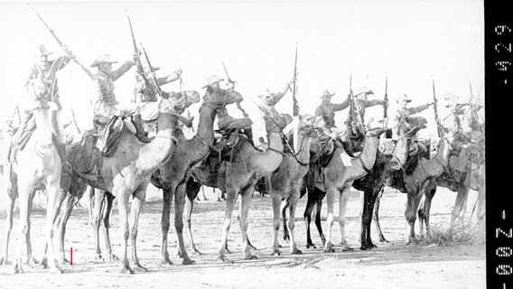 Historisches Schwarz-Weiß-Foto: Mehrere Menschen mit Gewehren sitzen auf Dromedaren. © Screenshot 