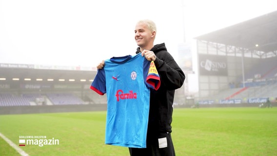 US-Fußballer John Tolkin hält ein Trikot des Fußballvereins Holstein Kiel in dessen Stadion hoch. © Screenshot 