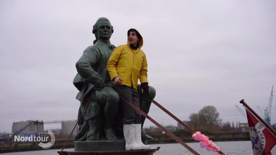 Ein Mann in gelbem Anorak steht zwischen den Beinen einer sitzenden Statue von Lessing auf einem kleineren Schiff das auf der Hamburger Elbe fährt. © Screenshot 