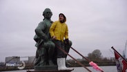 Ein Mann in gelbem Anorak steht zwischen den Beinen einer sitzenden Statue von Lessing auf einem kleineren Schiff das auf der Hamburger Elbe fährt. © Screenshot 