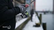 Eine Person hält eine Angel in der Hand an einem Kanal im Winter. © Screenshot 