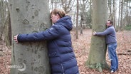 Eine Frau und ein Mann umarmen jeweils einen Baum in einem Wald. © Screenshot 