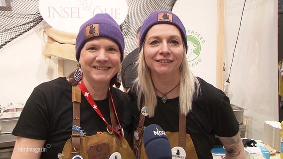 Zwei Frauen mit lilanen Beanies schauen freudig in die Kamera auf einer Landwirtschaftsmesse. © Screenshot 