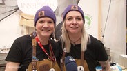 Zwei Frauen mit lilanen Beanies schauen freudig in die Kamera auf einer Landwirtschaftsmesse. © Screenshot 