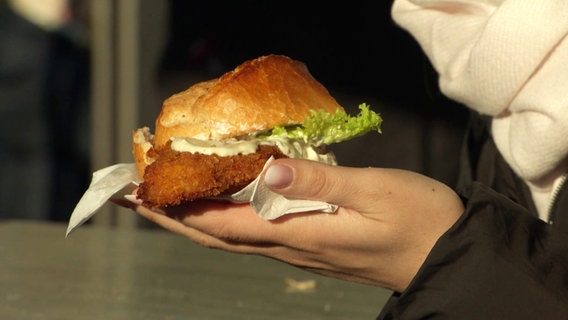 Eine Frau hält ein Brötchen mit etwas Frittiertem und Soße in der Hand. © Screenshot 