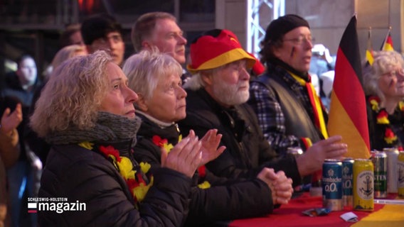 Personen in Deutschland-Outfit schauen auf einen Bildschirm. © Screenshot 