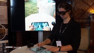 Eine Frau mit VR-Brille steht vor einem Bildschirm. © Screenshot 