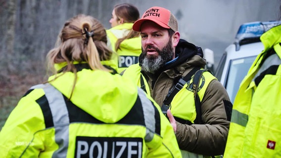 Polizeibeamte in Warnwesten sprechen in einem Wald mit Jägern. © Screenshot 