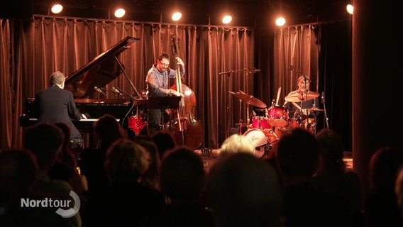 Ein Jazztrio spielt auf einer kleineren Bühne vor einem Bar-Publikum. © Screenshot 