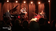 Ein Jazztrio spielt auf einer kleineren Bühne vor einem Bar-Publikum. © Screenshot 