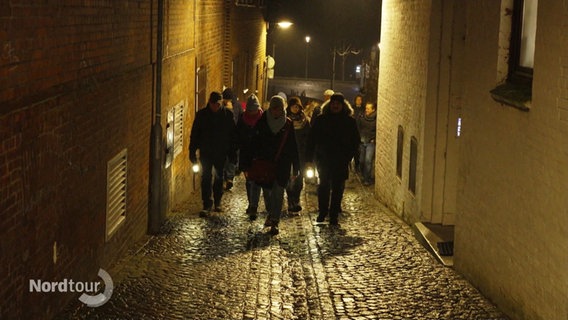 Eine Gruppe von Menschen geht bei Nacht mit Laternen durch eine enge Gasse. © Screenshot 