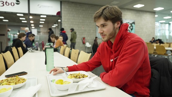 Ein Student sitzt an einem langen Tisch in einer Mensa, vor ihm ein Tablet mit Essen. © Screenshot 