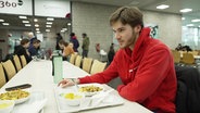 Ein Student sitzt an einem langen Tisch in einer Mensa, vor ihm ein Tablet mit Essen. © Screenshot 