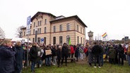 Mehrere Menschen haben sich auf einer Wiese vor einem Has zu einer Gegen-Demonstration zusammengefunden. © Screenshot 