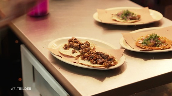 Auf einem Tisch stehen mehrere Teller mit Tacos. © Screenshot 