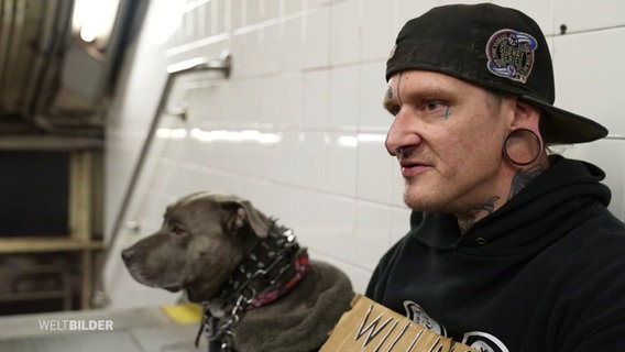 Ein mann mit einem großen Ohrtunnel und einem Hund sitzt auf dem Boden neben einer Treppe in einer U-Bahn-Station. © Screenshot 