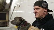 Ein mann mit einem großen Ohrtunnel und einem Hund sitzt auf dem Boden neben einer Treppe in einer U-Bahn-Station. © Screenshot 