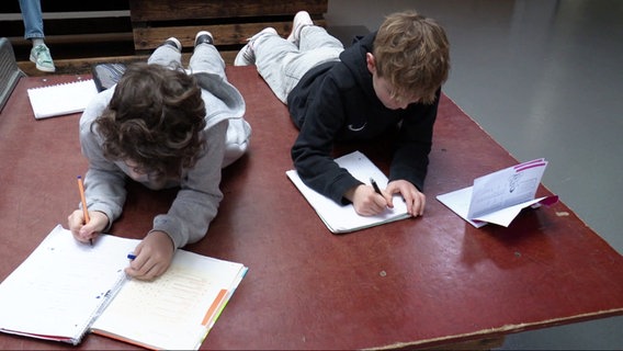 Zwei Kinder liegen auf einem Tisch in  einem Klassenraum und machen Schulaufgaben. © Screenshot 