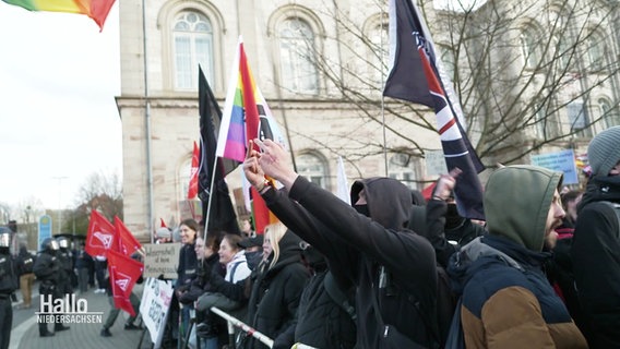 Demonstranten stehen hinter einer Absperrung. Über ihnen sind verschiedene Flaggen, darunter die Regenbogenflagge. Ein Demonstrant streckt die Mittelfinger empor. © Screenshot 