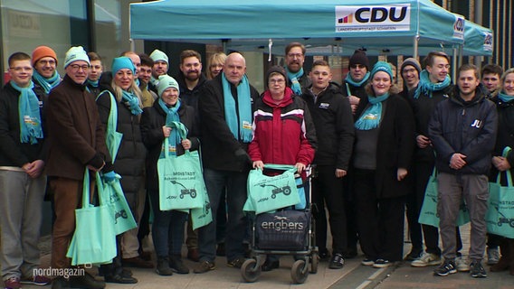 Mehrere CDU-Mitglieder posieren für ein Foto vor einem CDU-Infostand. Einige tragen einen Beutel, der im Türkiston der CDU gehalten ist. © Screenshot 