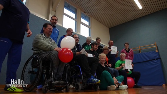 Eine Gruppe von Menschen posiert in einer Turnhalle für ein Foto. Ein Mann im Rollstuhl hält jubelnd zwei Luftballons, anderen halten eine Urkunde empor. © Screenshot 