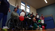 Eine Gruppe von Menschen posiert in einer Turnhalle für ein Foto. Ein Mann im Rollstuhl hält jubelnd zwei Luftballons, anderen halten eine Urkunde empor. © Screenshot 