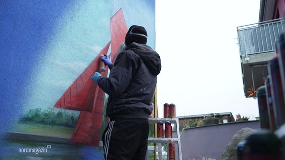 Mathias Oppen in schwarzer Kleidung sprayt ein Graffito an eine Hauswand. Zu erkennen ist eine Seenlandschaft. Er arbeitet gerade an roten Segeln. © Screenshot 