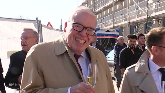 Milliardär Klaus-Michael Kühne lächelt mit einem Sektglas in der Hand in die Kamera. © Screenshot 