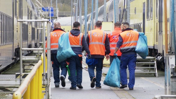 Fünf Männer mit orangen Westen und Mülltüten laufen an einem Zug entlang. © Screenshot 