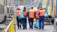 Fünf Männer mit orangen Westen und Mülltüten laufen an einem Zug entlang. © Screenshot 