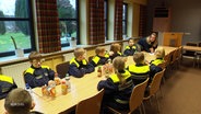 Die Kinderfeuerwehr in Marne sitzt in Arbeitskleidung an einem Tisch. © Screenshot 