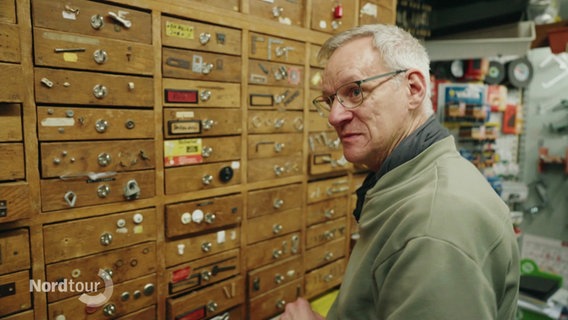 Ein Mann Steht vor einem Schrank mit zahlreichen kleinen Schubladen. © Screenshot 