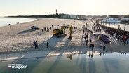 Eine Luftaufnahme zeigt, wie der Pottwal-Kadaver am Strand von Hörnum geborgen wird. Ein Bagger und Fachkräfte sind im Einsatz, dazu eine Menge Schaulustiger. © Screenshot 