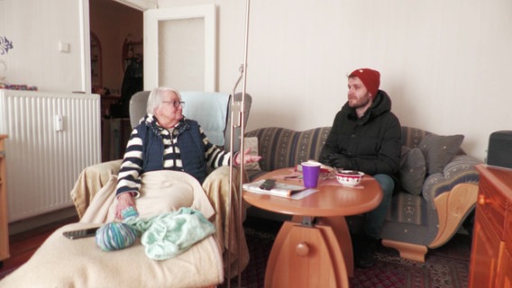 Ein Mann sitzt mit Mütze und Jacke auf einem Sofa. © Screenshot 