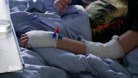 Ein Kinderarm mit Infusionen und Verband liegt auf einer Bettdecke eines Krankenhausbettes. © Screenshot 