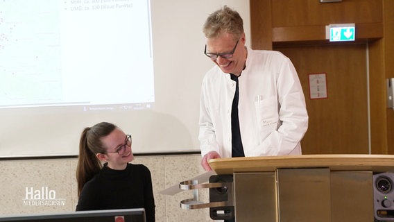 Prof. Dr. Nils Schneider von der Medizinischen Hochschule Hannover spricht im weißen Kittel im Hörsaal mit einer Studierenden. © Screenshot 