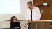 Prof. Dr. Nils Schneider von der Medizinischen Hochschule Hannover spricht im weißen Kittel im Hörsaal mit einer Studierenden. © Screenshot 