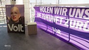 Ein Plakat und ein Banner der Volt-Partei stehen in einem Raum. © Screenshot 