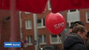 Ein roter Luftballon mit "SPD"-Aufdruck schwebt unter einem roten Pavillon. Im Hintergrund unscharf Menschen und Wohnhäuser. © Screenshot 